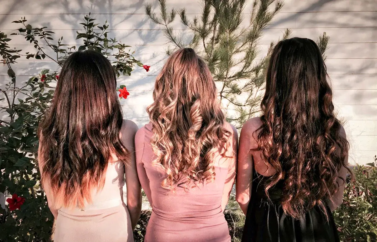 Tres mujeres de espaldas con melenas largas de rizos marcados, dos castañas y una rubia rosada, ante un seto con flores